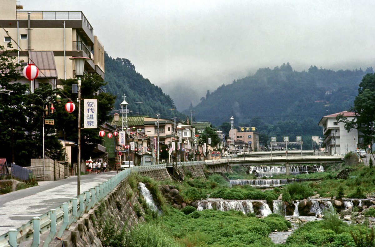 A riverside street with paper lanterns, small weirs in a green river and forested hills in mist, photographed in 1971