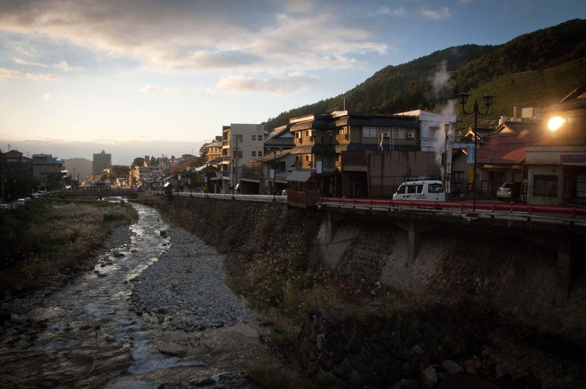 A shallow stony river running between stone embankments, with wooden and concrete buildings along the bank and wooded hills behind, late in the day