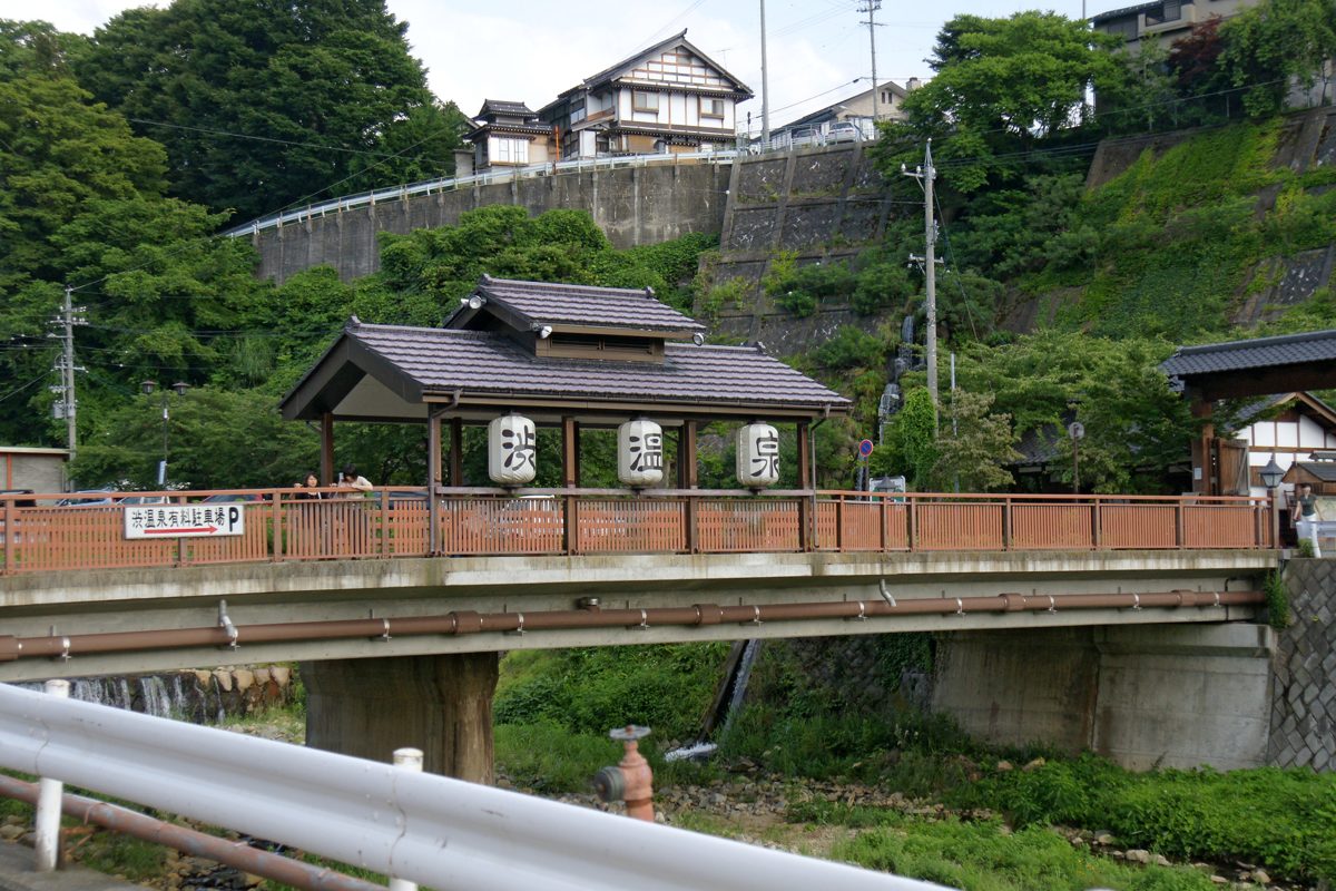 A roofed bridge with white lanterns over a river among green hills in Shibu Onsen