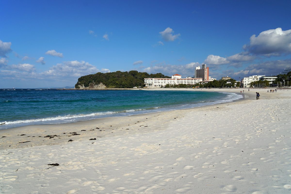 White sand beach with blue sea, a wooded headland and hotels in the distance on a sunny day