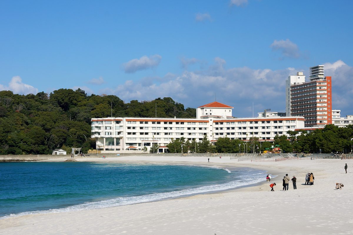 A curved white sand beach with blue sea, green hills and hotel buildings under a blue sky