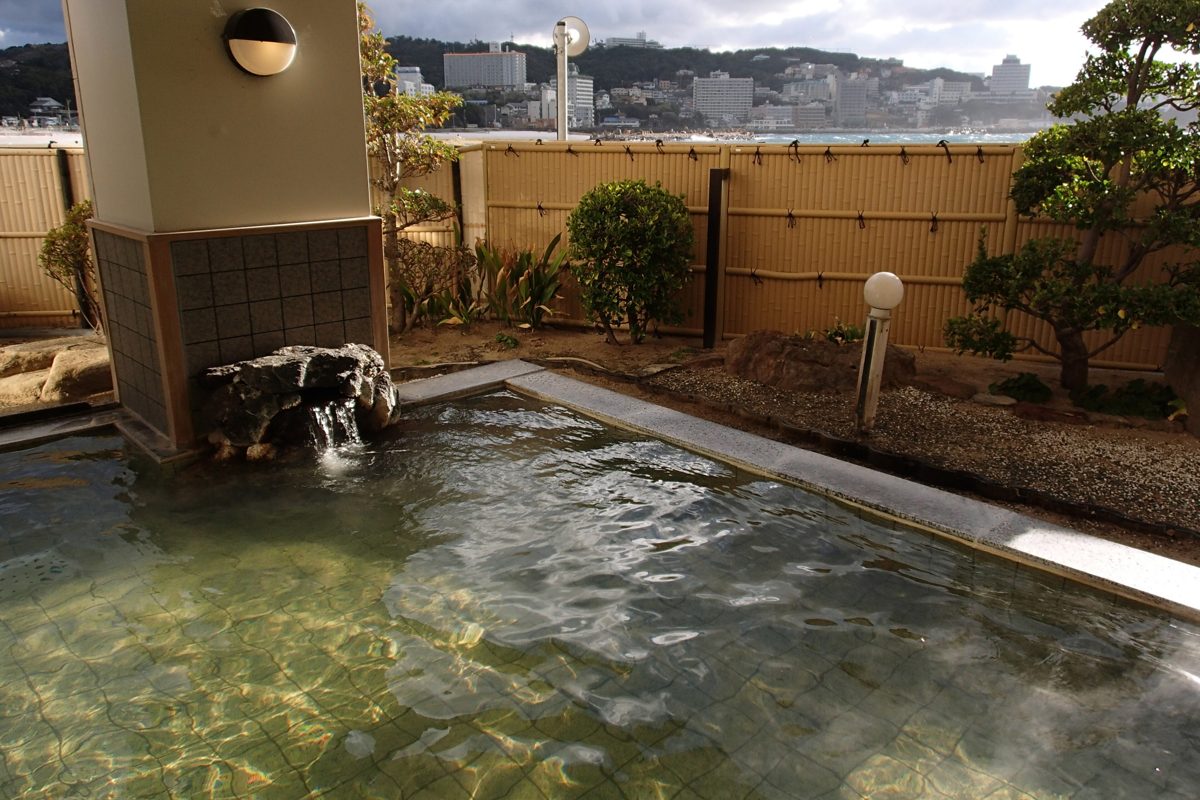 An open-air hot-spring pool with a small rock spout, a bamboo fence and hotels across the bay