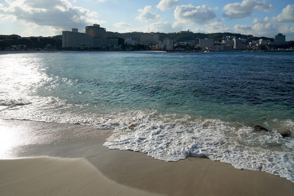 Waves washing onto white sand at Shirarahama Beach on a sunny day, with buildings along the far shore
