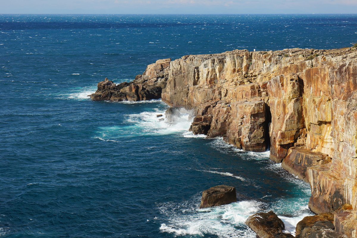 Tall sandstone cliffs dropping into a blue sea with white waves on a sunny day