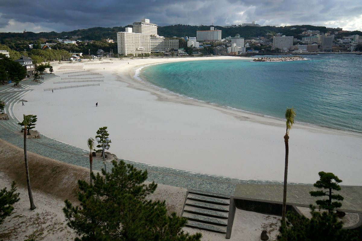 A curved white sand beach with turquoise water, palm trees and hotels behind it