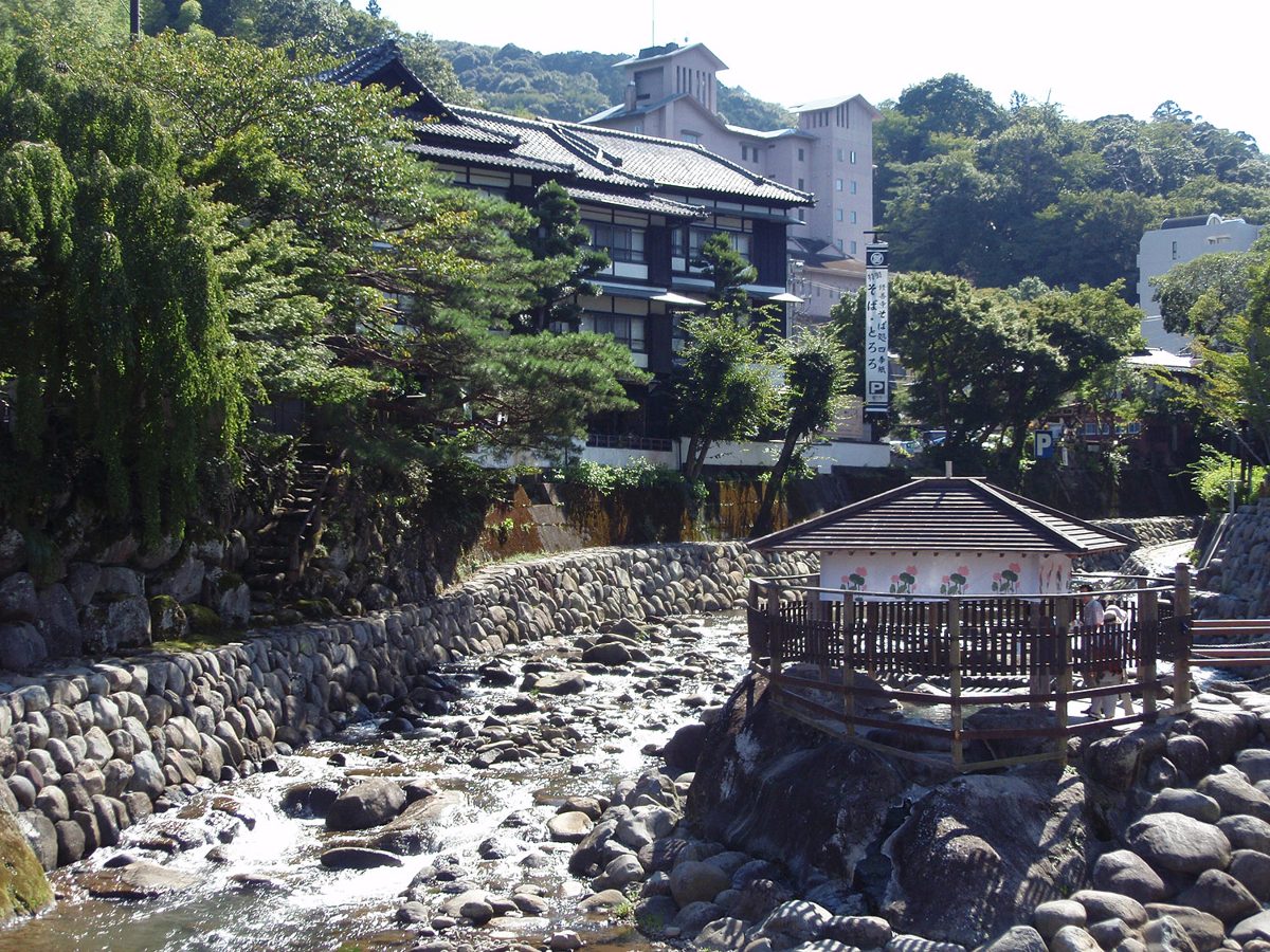 A small roofed structure on rocks in a stony river, with stone embankments and wooden buildings behind