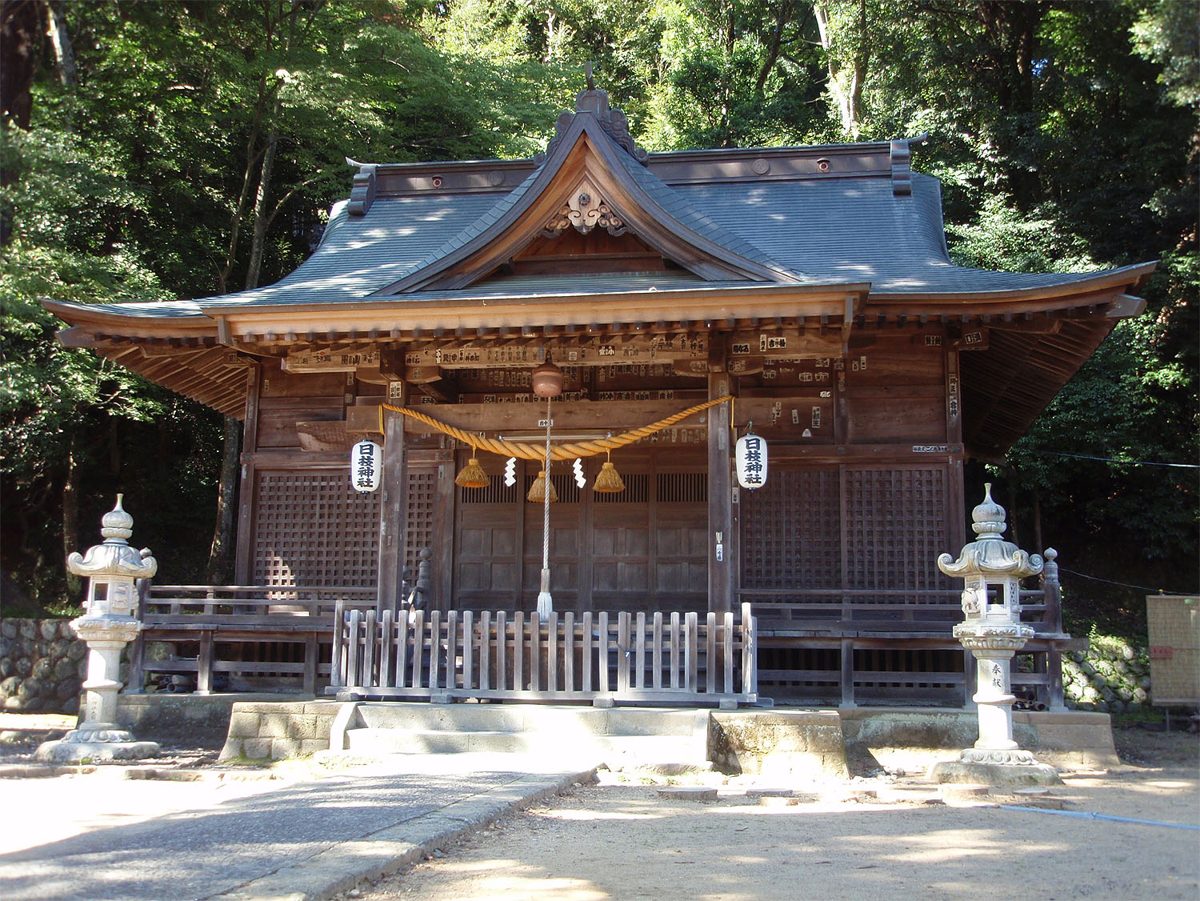 A wooden Shinto shrine hall with stone lanterns in front, surrounded by trees