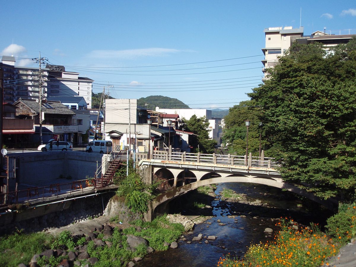 An old concrete arch bridge over a shallow rocky river, with town buildings and green hills under a blue sky