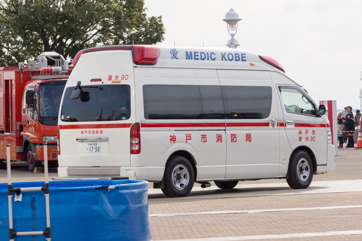 A white Kobe City Fire Bureau ambulance with red stripes parked next to a red fire truck
