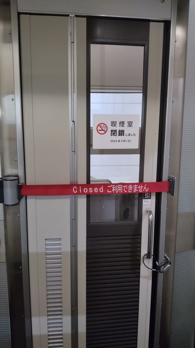 A glass-paneled smoking room door blocked by a red Closed belt, with a sign saying the smoking room closed on March 1, 2024