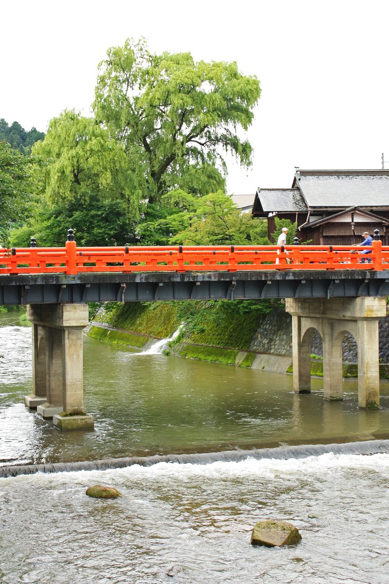 A red-railed bridge over a shallow river with willow trees behind it