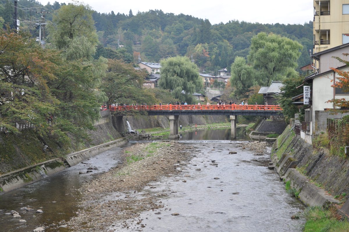 A red bridge over the shallow Miya River in Takayama, with trees and houses on both banks