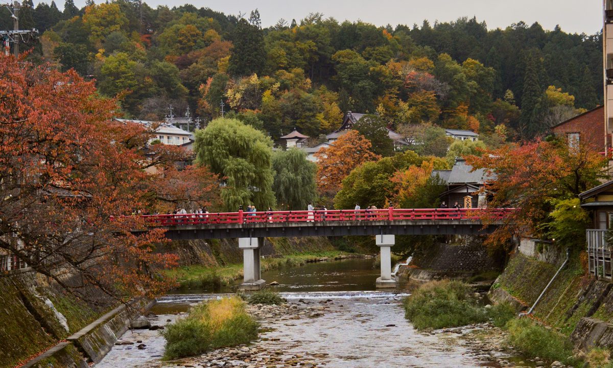 A red bridge over a shallow river with autumn trees and wooded hills in Takayama