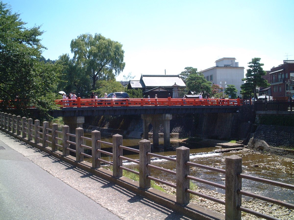 A red bridge over a shallow river on a sunny day, with trees and low buildings behind