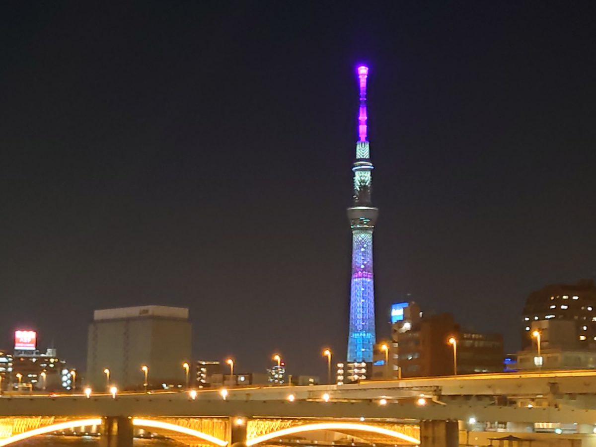 Tokyo Skytree lit in blue and purple at night, behind a lit bridge over a river