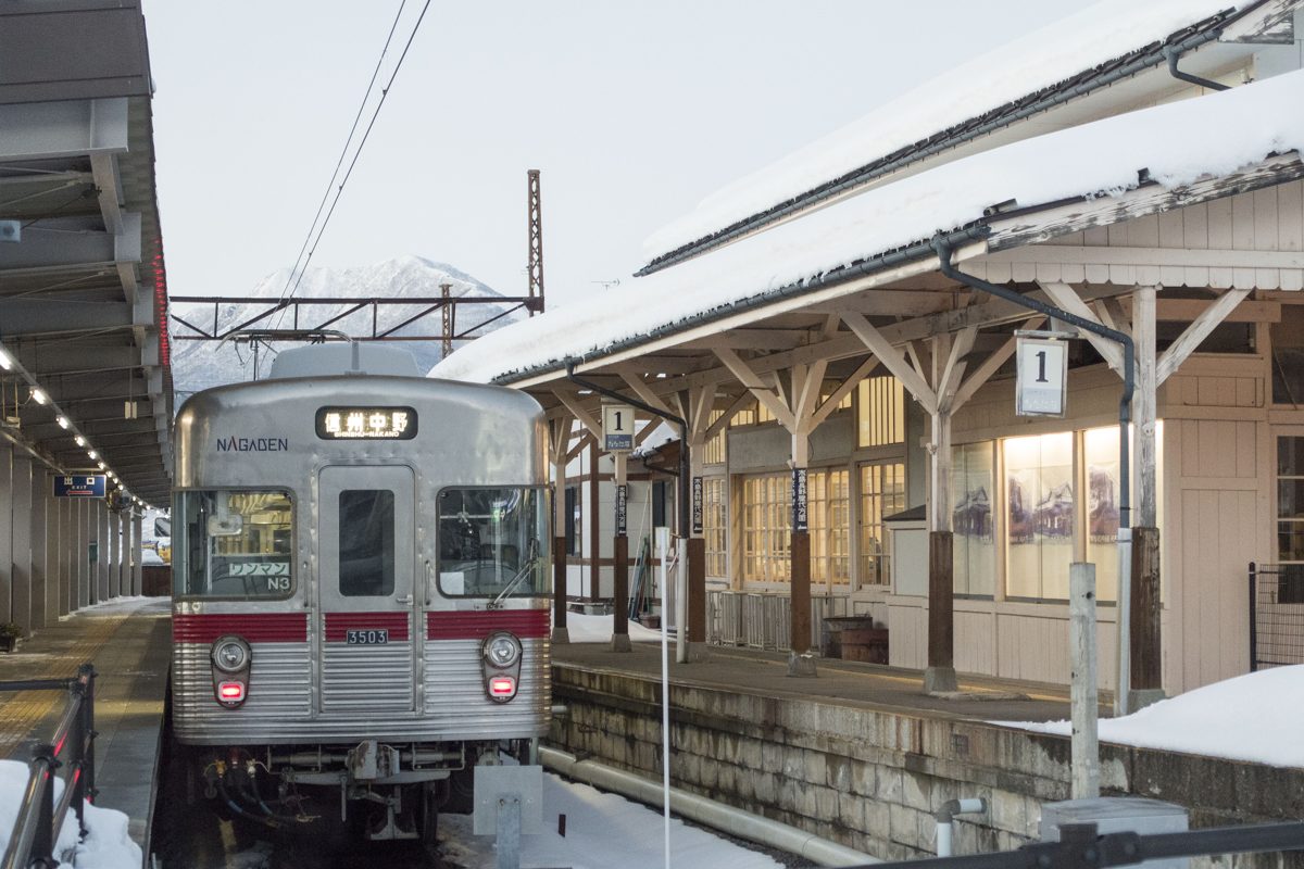 A silver Nagano Electric Railway train with a red stripe at a snowy platform under a wooden canopy
