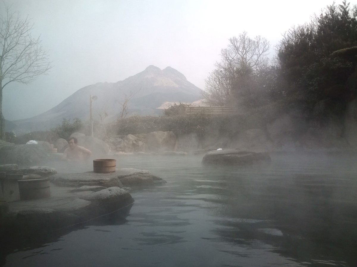 A steaming outdoor rock bath with Mount Yufu faintly visible behind trees