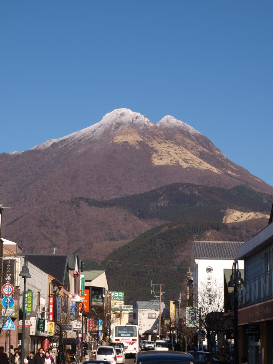 A street with shops and signs in Yufuin, with snow-capped Mount Yufu behind