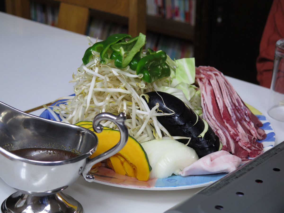 A plate of raw lamb, bean sprouts, cabbage and other vegetables with a jug of sauce