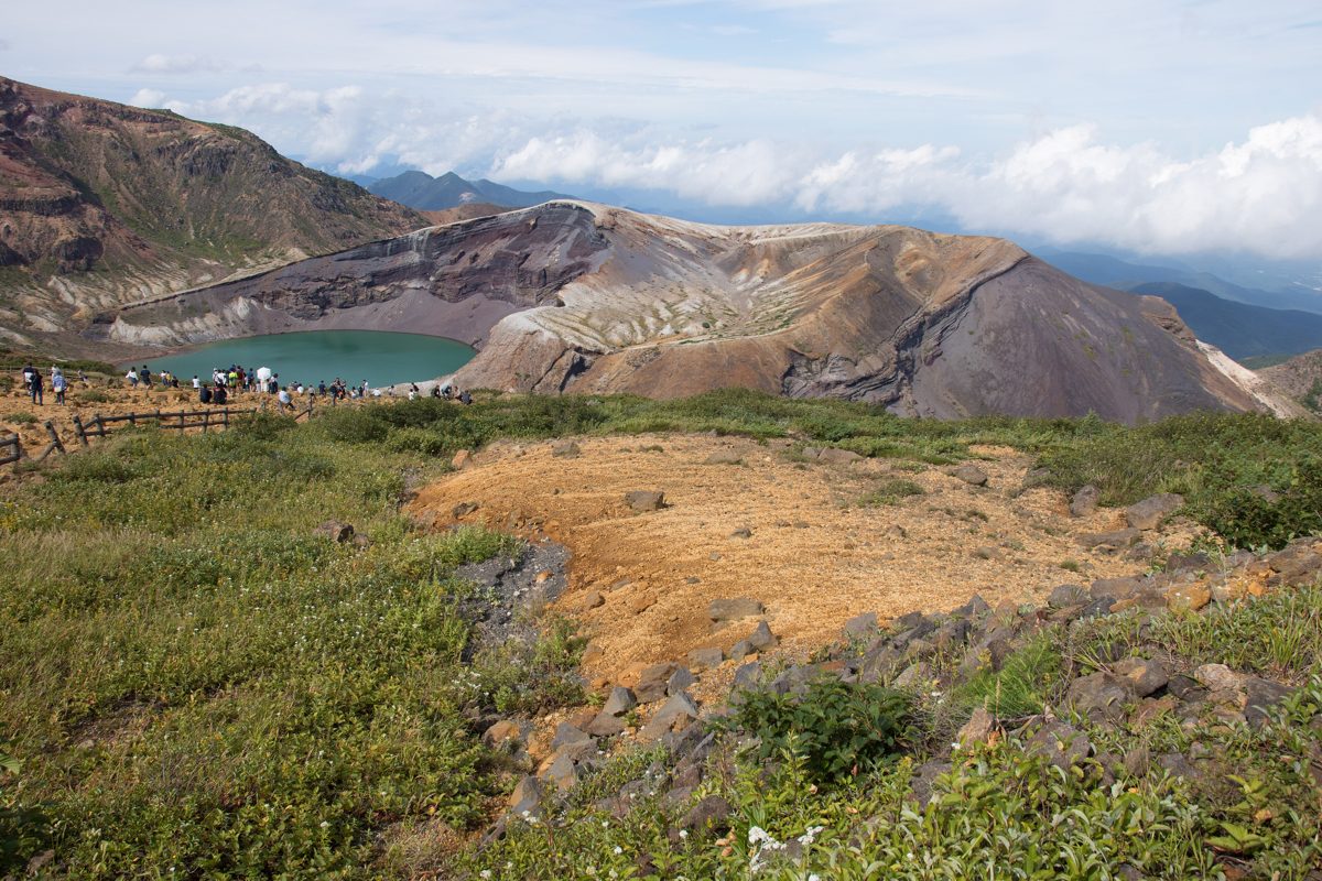 A green crater lake in a brown volcanic crater under a cloudy sky