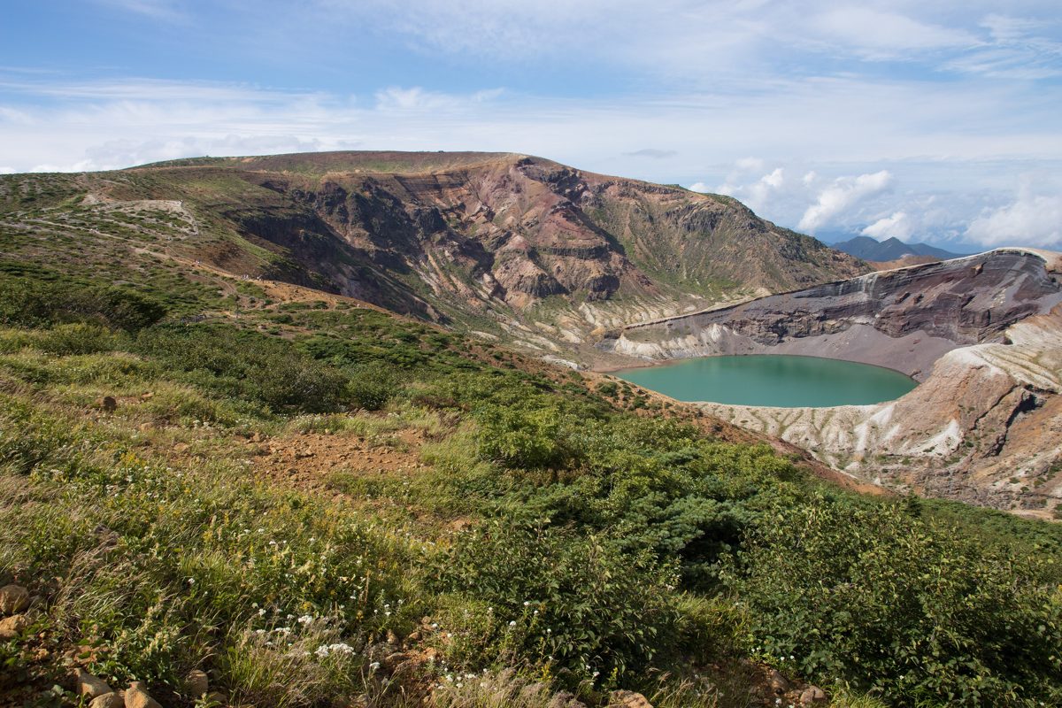 A green crater lake inside a brown volcanic crater on a sunny day, with grassy slopes in front